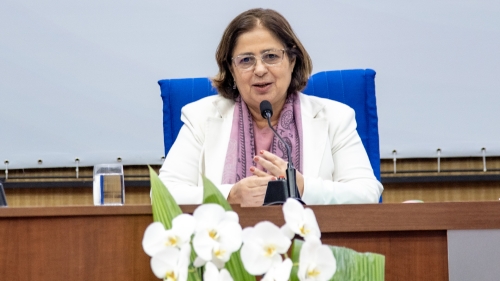 A woman in a white blazer speaks at a podium, with a microphone and a floral arrangement in front of her.